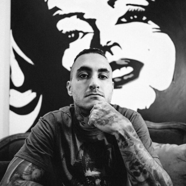 Homem com os braços tatuados com a mão no queixo assentado em um estúdio de tatuagem com uma pintura ao fundo em preto e branco
