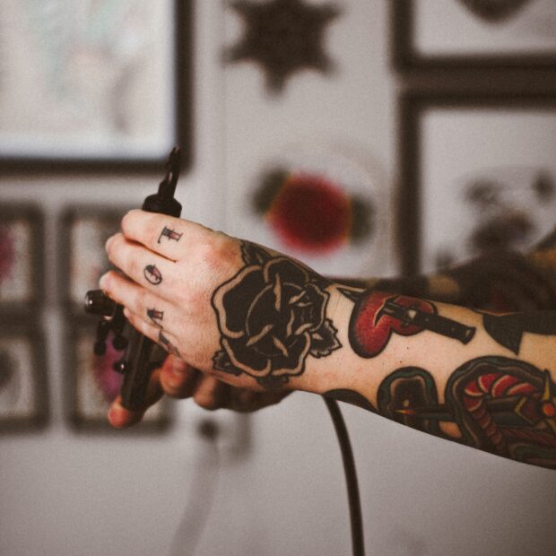 Braco-tatuado-segurando-maquina-de-tatuagem-com-quadros-ao-fundo