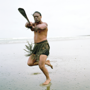 homem-com-saia-de-folhas-com-remo-apontando-para-câmera-rosto-tatuado-maori-mar-ao-fundo